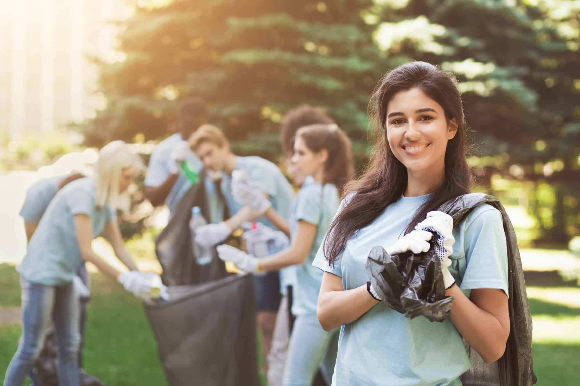A volunteer smiles at the camera with a trash bag slung around their shoulder while others in the background help clean up a park.